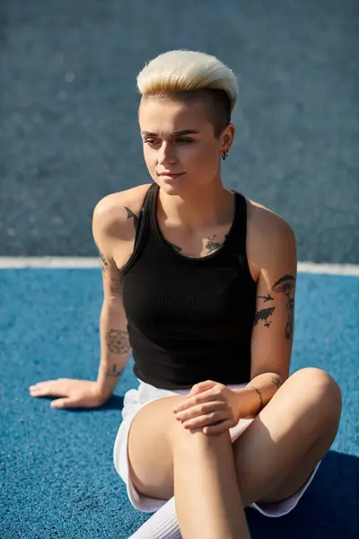 Una joven con el pelo corto y tatuajes sentada con las piernas cruzadas en el suelo en un entorno al aire libre sereno. - foto de stock