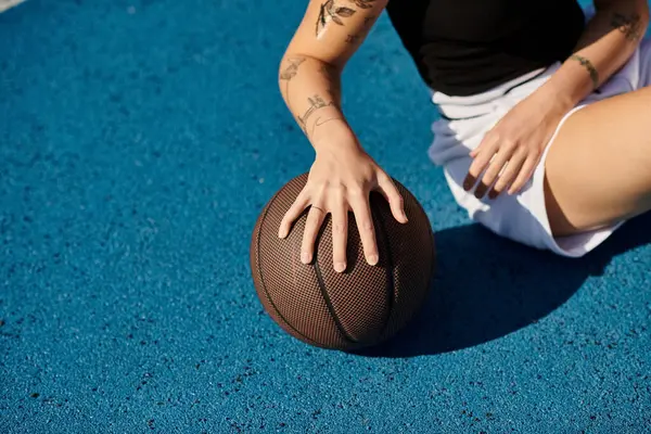 Молода жінка з татуюваннями сидить на землі, тримаючи баскетбол, виключаючи ауру рішучості та атлетизму. — стокове фото