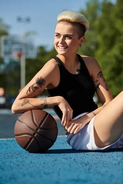 Una joven con el pelo corto y tatuajes se sienta en el suelo, sosteniendo una pelota de baloncesto en un entorno urbano al aire libre. - foto de stock