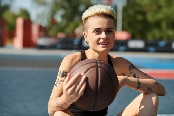 Una giovane donna con i capelli corti e tatuaggi si siede a terra, cullando una pallacanestro con uno sguardo determinato. — Foto stock