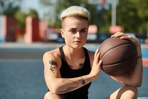 Une jeune femme aux cheveux courts et tatouée assise sur le sol, tenant un ballon de basket, perdue dans la pensée. — Photo de stock