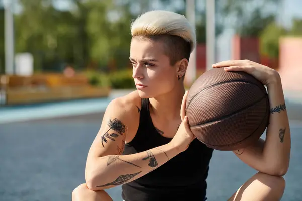 Una joven con el pelo corto y tatuajes se sienta en el suelo, profundamente pensada mientras sostiene una pelota de baloncesto en sus manos. - foto de stock