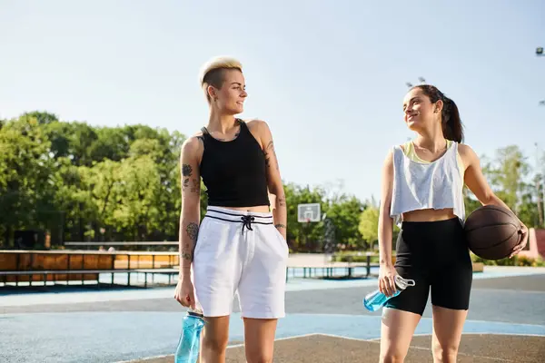 Two young women, friends, stand together on a basketball court, showcasing their sporty skills with a backdrop of a sunny summer day. — Stock Photo