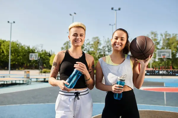 Dos mujeres jóvenes, sosteniendo botellas de agua, se preparan para jugar al baloncesto al aire libre en un día soleado en verano. - foto de stock