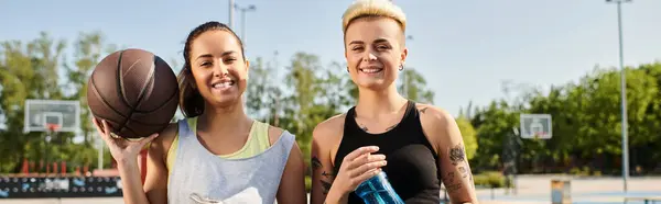 Dos mujeres jóvenes, atléticas y en ropa deportiva, se paran juntas al aire libre, sosteniendo una pelota de baloncesto. - foto de stock