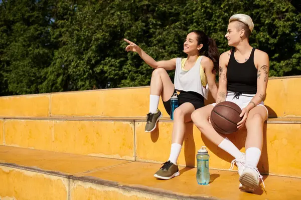 Две молодые женщины, спортивные друзья, сидят вместе после игры в баскетбол на открытом воздухе в летний день. — стоковое фото