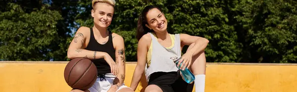Two young women in athletic wear are sitting on top of a basketball court, chatting and enjoying a relaxing break after playing a game. — Stock Photo