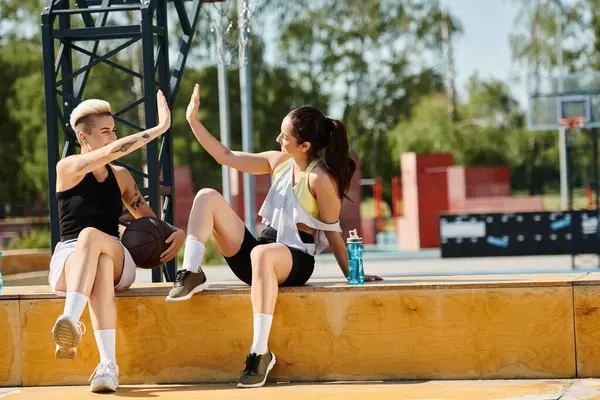 Two young women, athletic friends, sitting on a ledge with their arms in the air in a moment of celebration after playing basketball outdoors. — Stock Photo