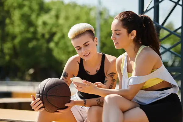 Two young women sitting on a bench outdoors, engrossed in a cell phone screen. — Stock Photo