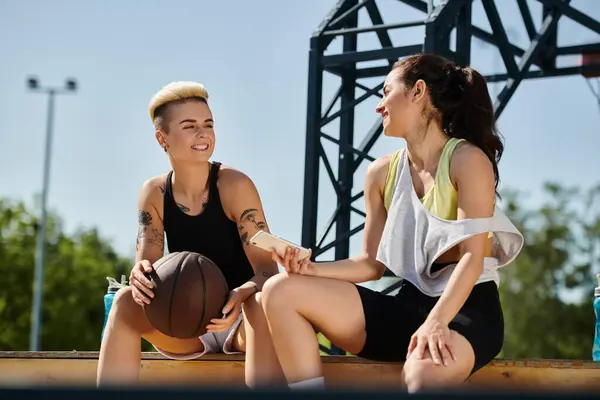 Deux jeunes femmes, athlétiques et pleines de vigueur, profitent d'une journée ensoleillée à l'extérieur alors qu'elles s'assoient par terre avec un ballon de basket. — Photo de stock