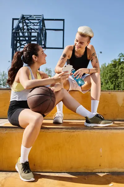 Dos jóvenes atléticas, amigas, se sientan en las escaleras, charlando y compartiendo historias durante un descanso de jugar al baloncesto al aire libre en verano. - foto de stock