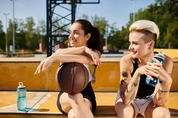 Dos mujeres jóvenes con una pelota de baloncesto sentadas en un banco, que participan en un partido amistoso de baloncesto al aire libre. - foto de stock