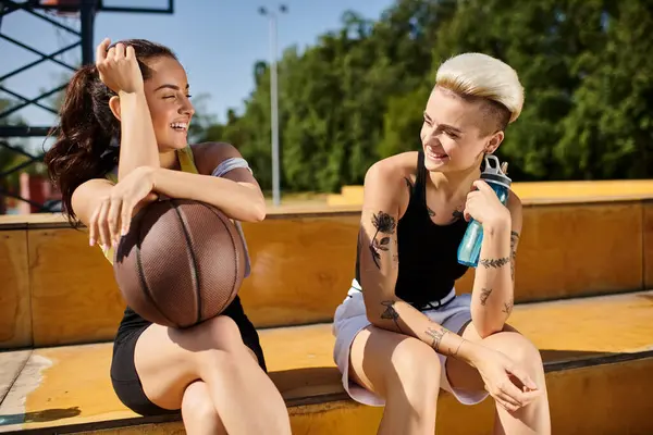 Две молодые атлетичные женщины сидят бок о бок, делая перерыв от игры в баскетбол на открытом воздухе в солнечный летний день. — стоковое фото