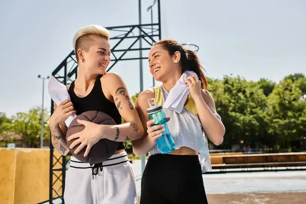 Dos mujeres jóvenes, amigos y atletas, se paran con confianza una al lado de la otra en una cancha de baloncesto al aire libre en el verano. - foto de stock
