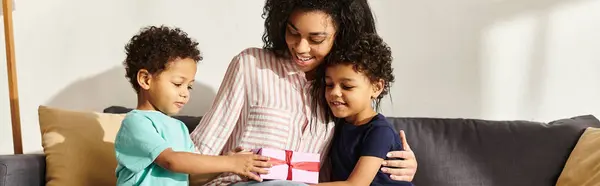 Hermosa alegre afroamericana mujer recibiendo regalo de sus adorables hijos en el día de la madre - foto de stock