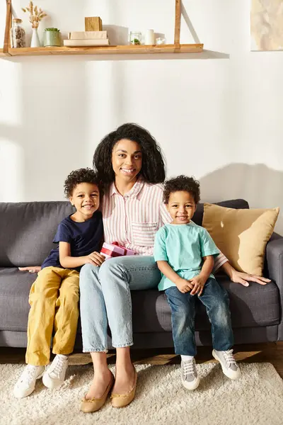 Alegre afroamericana madre sosteniendo presente y sentado en sofá con sus hijos, mirando a la cámara - foto de stock