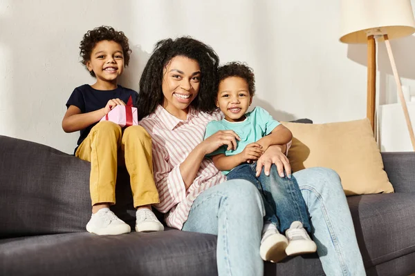 Familia afroamericana alegre de tres sonriendo alegremente a la cámara, presente en la mano, Día de las Madres - foto de stock