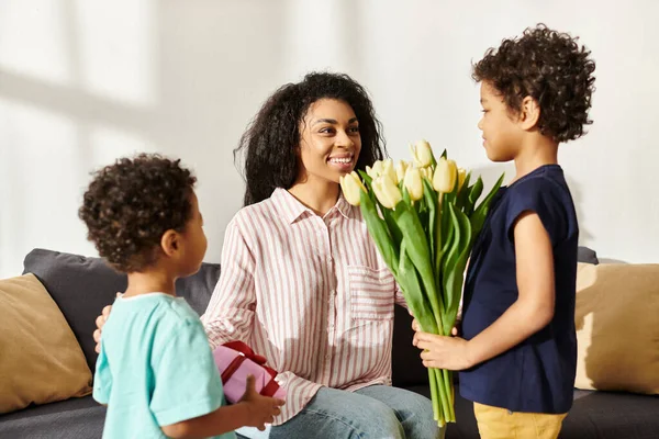 Belle femme afro-américaine joyeux recevant un cadeau et des tulipes de ses petits fils, fête des mères — Photo de stock