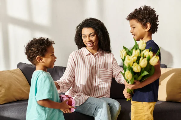 Joyeuse femme afro-américaine recevant un cadeau et des tulipes de ses petits fils aimants, fête des mères — Photo de stock