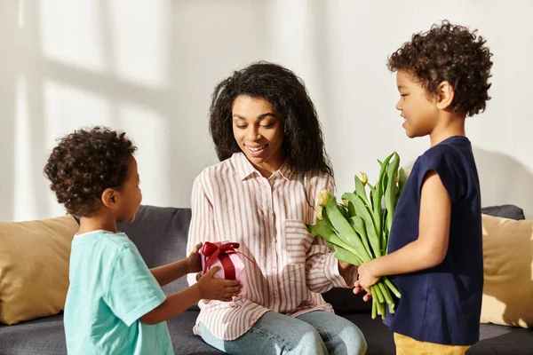Joyous african american woman receiving gift and tulips from her adorable sons on Mothers day — Stock Photo