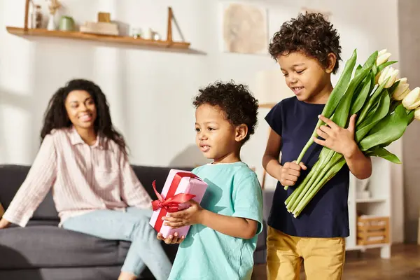 Focus on merry african american boys with tulips and gift with their blurred mother on backdrop — Stock Photo