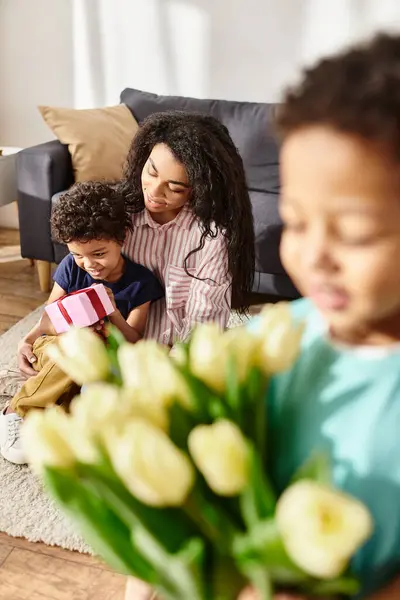 Se concentrer sur joyeux afro-américaine mère avec fils tenant cadeau près de son autre fils flou avec des tulipes — Photo de stock