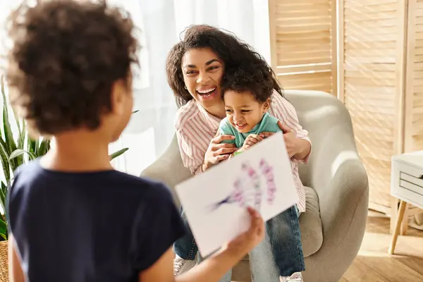 Jolly african american woman receiving postcard from her son while holding his brother, Mothers day — Stock Photo