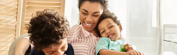 Alegre afroamericana madre sonriendo y abrazando calurosamente a sus adorables y alegres hijos, bandera - foto de stock
