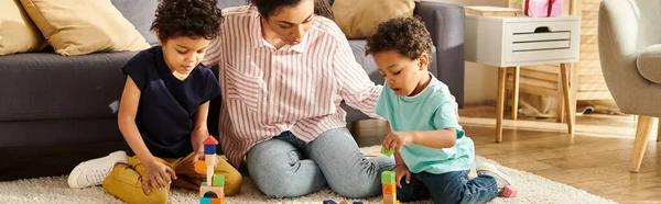 Hermosa mujer afroamericana en ropa de casa jugando con sus adorables hijos en casa, pancarta - foto de stock