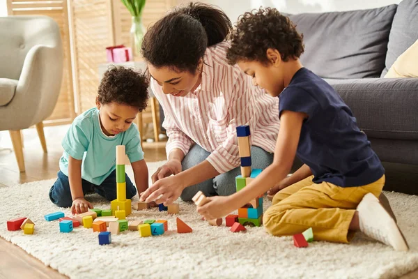 Alegre atraente afro-americana mãe em roupas aconchegantes brincando com brinquedos com seus pequenos filhos bonitos — Fotografia de Stock