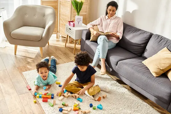 Alegre afro-americana mãe olhando para seus filhos brincando com brinquedos enquanto ela lendo livro — Fotografia de Stock