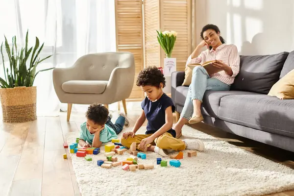 Alegre afro-americana mãe em casa assistindo seus filhos jogar enquanto ela lendo livro — Fotografia de Stock