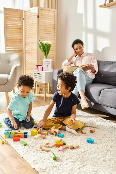 Fröhliche afrikanisch-amerikanische Frau in Hauskleidung liest Buch und schaut ihren Kindern beim Spielen mit Spielzeug zu — Stockfoto