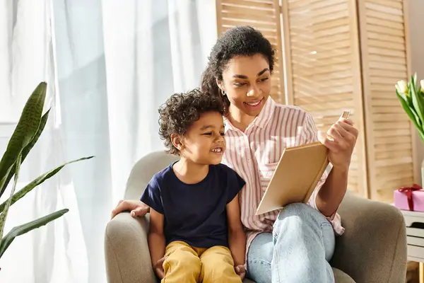 Hermosa mujer afroamericana alegre y su adorable hijo leyendo libro juntos mientras están en casa - foto de stock