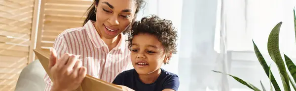 Alegre atractiva mujer afroamericana en traje acogedor libro de lectura a su pequeño hijo en casa, pancarta - foto de stock