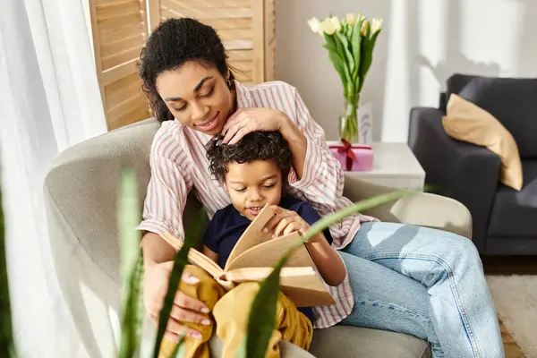 Alegre atractiva africana americana mujer en traje acogedor libro de lectura con su pequeño hijo alegre - foto de stock