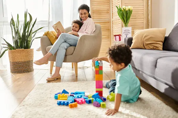 Pequeño chico afroamericano lindo jugando con juguetes con su madre y su hermano leyendo en el telón de fondo - foto de stock