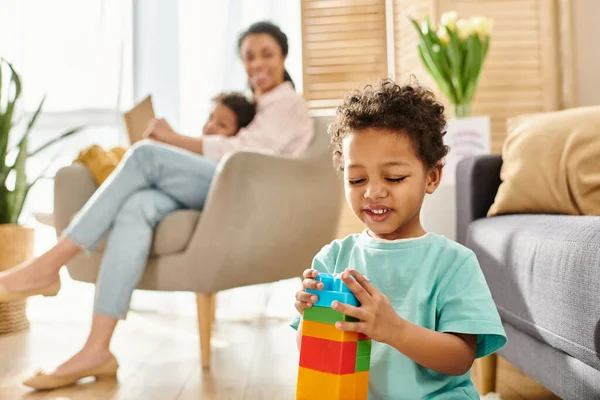 Enfoque en el niño afroamericano jugando con juguetes con su borrosa mamá y su hermano en el telón de fondo - foto de stock