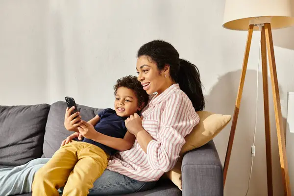 Atractiva madre afroamericana alegre y su adorable hijo sentado en el sofá y mirando el teléfono - foto de stock