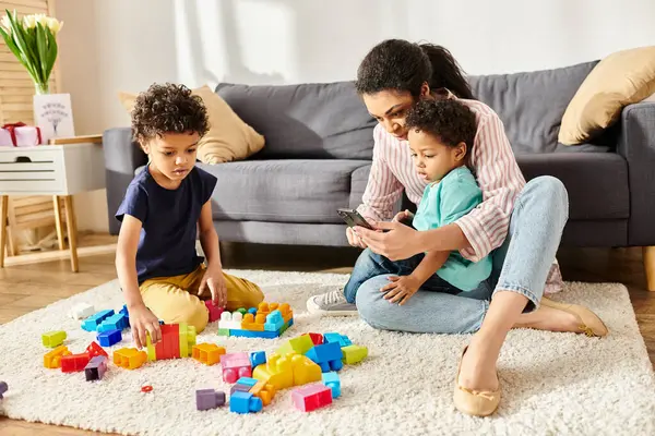 Hermosa madre afroamericana mirando el teléfono mientras sus hijos juegan con cubos en casa - foto de stock