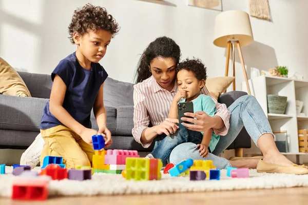 Mujer afroamericana guapa sosteniendo el teléfono mientras sus hijos pequeños juegan con cubos en casa - foto de stock