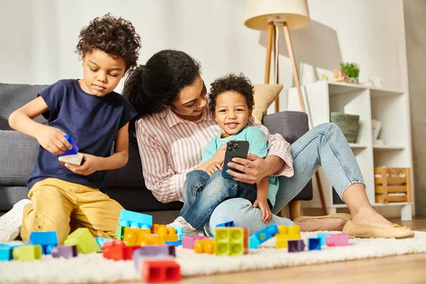 Freudige afrikanisch-amerikanische Mutter mit Telefon in der Hand, die Zeit mit ihren beiden entzückenden Söhnen verbringt — Stockfoto