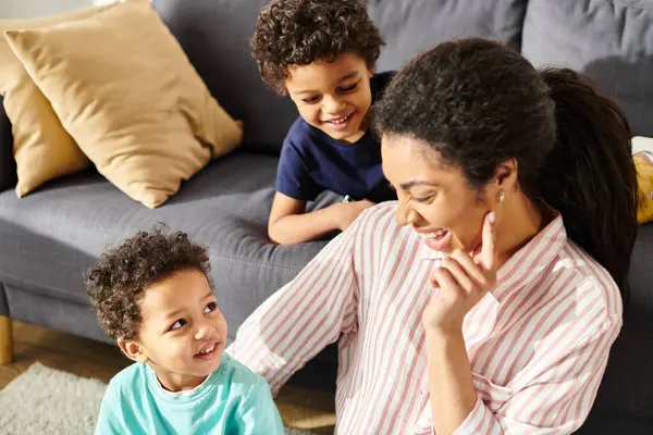 Fröhliche afrikanisch-amerikanische Mutter in Hauskleidung sitzt neben ihren kleinen Söhnen mit Laptop im Wohnzimmer — Stockfoto