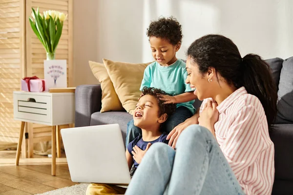 Joyeuse mère afro-américaine en tenue de maison regardant avec amour ses petits fils mignons avec ordinateur portable — Photo de stock