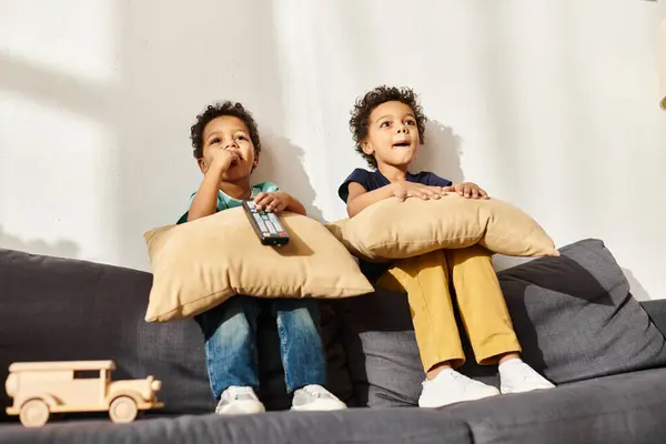 Joyful african american cute brothers sitting on sofa with pillows and watching TV in living room — Stock Photo