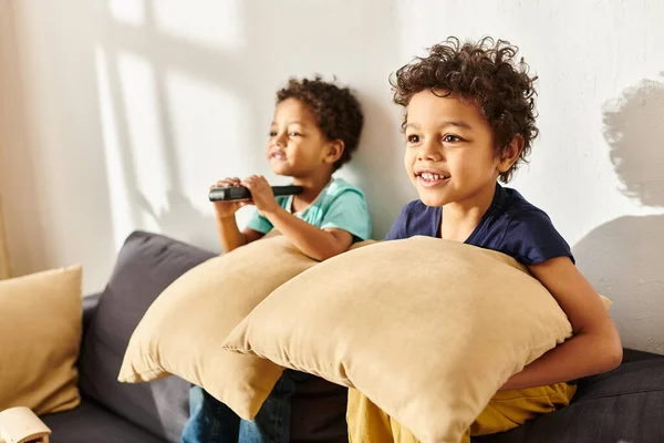 Foco em alegre menino americano africano com travesseiro assistindo TV ao lado de seu irmão adorável borrado — Fotografia de Stock