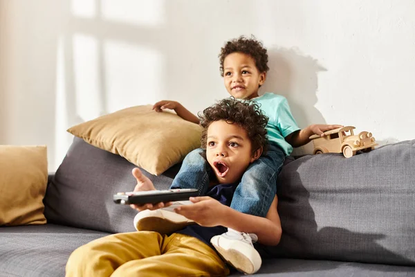 Chocado afro-americano menino segurando controle remoto e assistindo filmes com seu irmão bonito — Fotografia de Stock