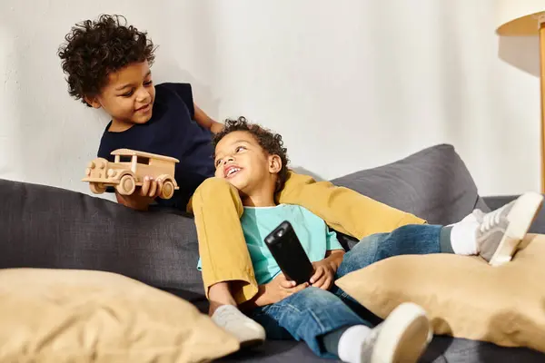 Радісні афроамериканські хлопчики сидять на дивані, граючи з іграшковим автомобілем і дивлячись один на одного — стокове фото