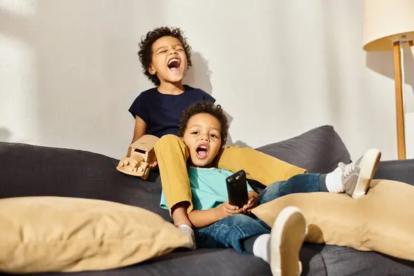 Веселые африканские маленькие братья сидят на диване с деревянной игрушечной машиной и пультом дистанционного управления — стоковое фото