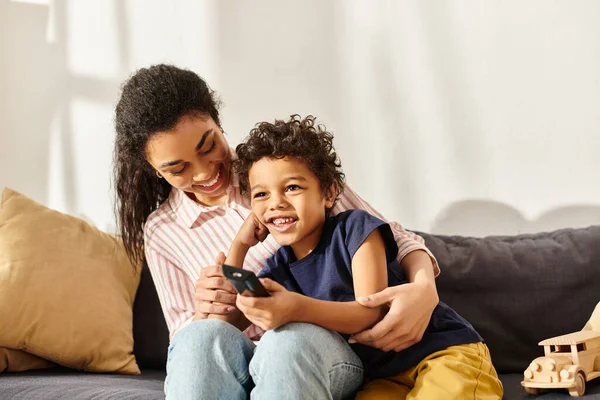 Joyeuse belle femme afro-américaine assise sur le canapé avec son adorable fils et regardant des films — Photo de stock